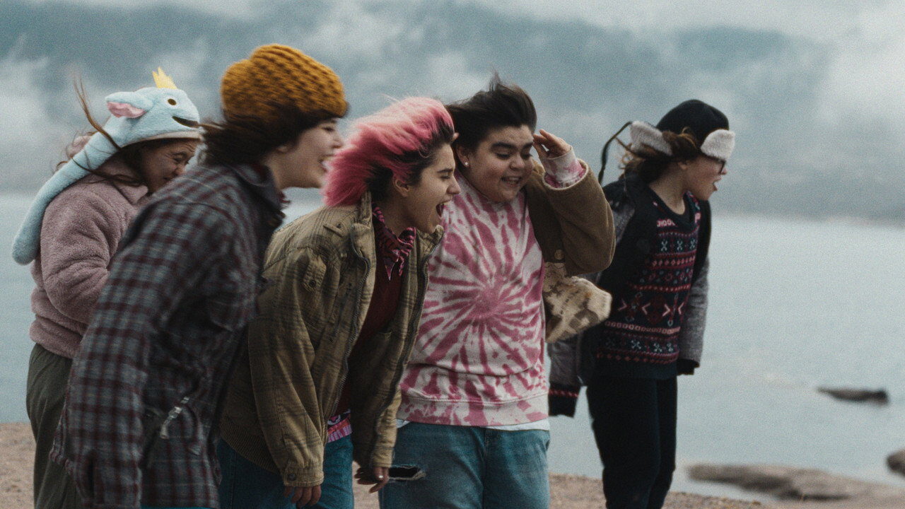 A group of teenagers in hats and jackets lean forward on a lakeshore under a grey sky